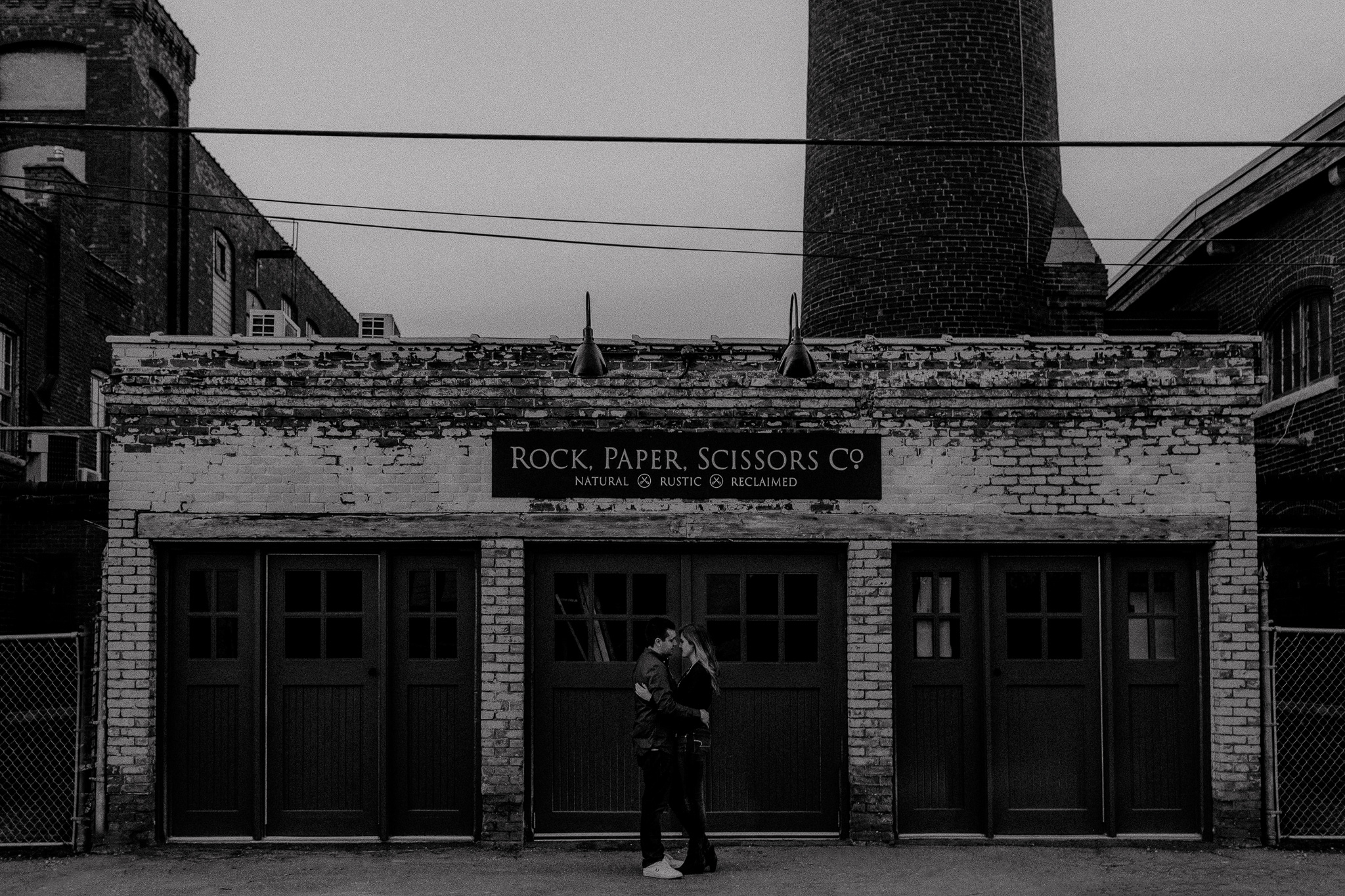 Engagement photo in front of Rock, Paper Scissors Co. sign at The Cotton Factory