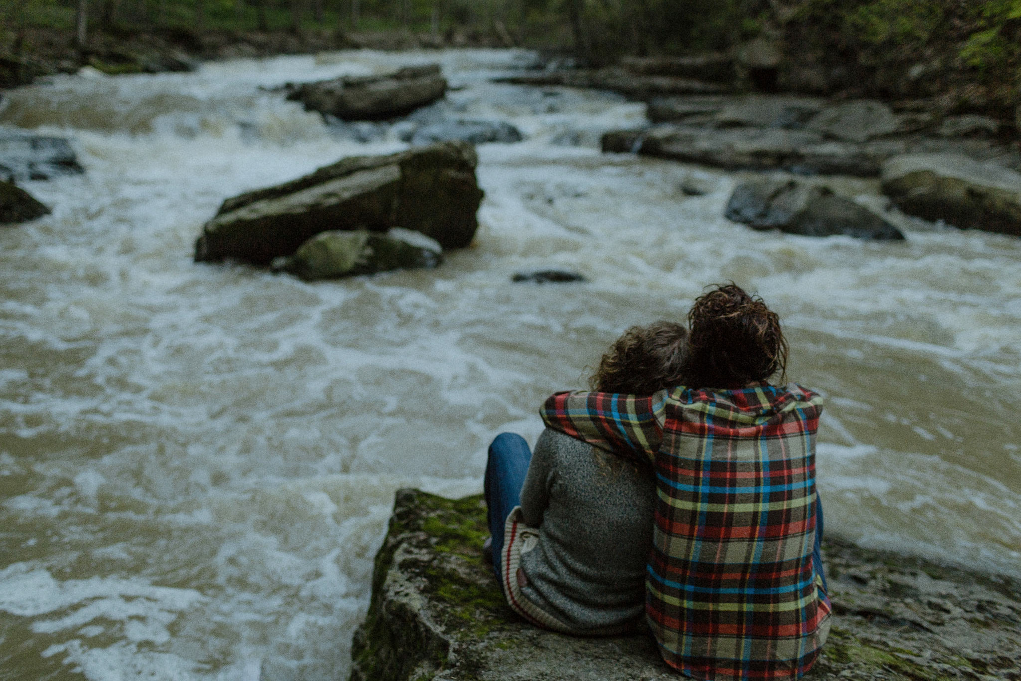 alternative rock couple sitting side by side on a mossy rock watching the water rapidly flow by